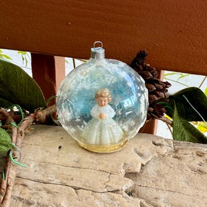 May include: A vintage Christmas ornament featuring a small figurine of a girl inside a clear glass globe. The globe has a silver cap and is decorated with blue and white accents. The figurine is wearing a white dress.