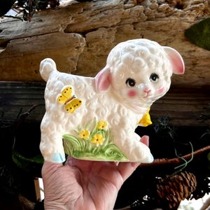 May include: A white ceramic lamb figurine with a textured curly coat, pink-tinted ears and cheeks, and large dark eyes. The lamb features a yellow butterfly on its side, a yellow neck accent, and a base decorated with green grass and yellow flowers.