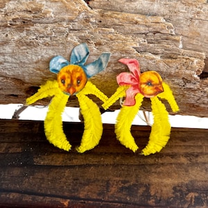 Puede incluir: Dos adornos de Pascua vintage con cuerpos de chenilla amarilla y caras de pollitos. Uno tiene un lazo azul, el otro un lazo rosa. Se colocan sobre un fondo de madera rústica.