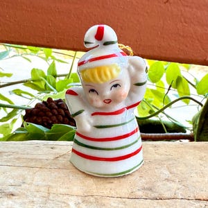 Puede incluir: Figura de cerámica vintage de un ángel sonriente con cabello rubio, vistiendo un atuendo blanco con rayas rojas y verdes. El ángel tiene las manos levantadas y lleva un sombrero a juego. La figura está sobre una superficie de madera.