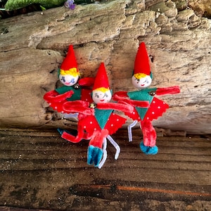 May include: Three red and green felt Christmas elf ornaments with pipe cleaner arms and legs. The elves have painted faces with yellow hair and black eyes.