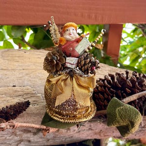 Puede incluir: Un adorno decorativo que presenta una pequeña figura con un gorro color jengibre y ropa roja, colocada sobre una base dorada texturizada. El adorno está decorado con piñas marrones, una cinta dorada y una pequeña caja de regalo plateada con borde rojo.