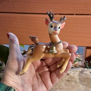 Puede incluir: Una figura de reno navideño vintage con un collar de campana. El reno es marrón con manchas blancas, una cara blanca y astas negras. La campana es de color dorado. La figura se sostiene en una mano.