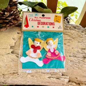 Op de afbeelding: Een pakket van twee vintage kerstengeldecoraties. De engelen hebben gouden vleugels, witte en roze jurken en rode sjaals. Op de verpakking staat "TIE-ON ALL PURPOSE Christmas DECORATIONS."