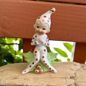 Pode incluir: Uma estatueta de elfo em cerâmica com base branca, decorada com bolinhas vermelhas e douradas. O elfo usa um chapéu pontudo com ponta dourada, está sentado sobre folhas de azevinho verdes e tem três bagas douradas na base. A figura tem traços pintados.