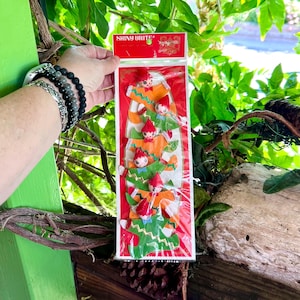 Op de afbeelding: Een pakketje vintage Shiny Brite kerstversiering. De versieringen zijn elfachtige figuren met rode hoeden, oranje jasjes en groene boomvormige lichamen. De verpakking heeft een rode achtergrond met het Shiny Brite logo bovenaan.