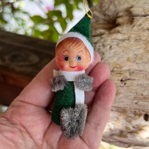 May include: A small elf ornament featuring a green felt hat and body, a white felt scarf, and grey fuzzy pipe cleaner accents for hands and feet. The elf has a painted face with rosy cheeks, blue eyes, and blonde hair, topped with a gold hanging string.