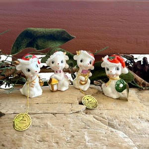 May include: Four white ceramic lamb figurines wearing red and green Christmas hats. Each lamb features gold-toned accents and holds a different object: a bell, a lantern, a saxophone, or a wreath. Each figurine has a gold string with a round tag.
