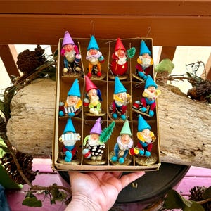 May include: A collection of twelve vintage gnome ornaments in a cardboard box. Each gnome has a different colored hat, including blue, red, purple, and green. The gnomes are dressed in colorful outfits and are holding various items.