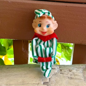 Puede incluir: Una pequeña figura de elfo con rostro pintado, ojos azules y mejillas sonrosadas. El elfo viste un conjunto de tela a rayas verdes y blancas con un gorro puntiagudo a juego y un cuello rojo sólido. La figura está sentada con los brazos cruzados.