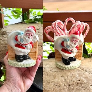 May include: A ceramic Santa Claus figurine with a red hat and coat, black boots, and a white beard. The figurine is holding a blue sack and is set against a brick wall. The base is green and the figurine is filled with candy canes.