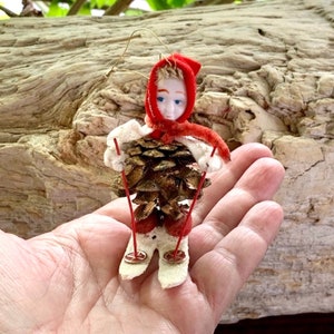 May include: A vintage Christmas ornament in the shape of a skier. The skier's head is a porcelain doll face with blue eyes and blonde hair, wearing a red hat and scarf. The body is a pine cone, with white felt arms and legs, and red skis.