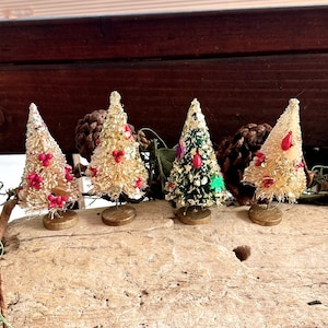 Puede incluir: Una colección de árboles de Navidad en miniatura en varios colores, incluyendo dorado y verde, adornados con bayas rojas y adornos. Cada árbol está montado sobre una pequeña base redonda, creando una exhibición festiva.