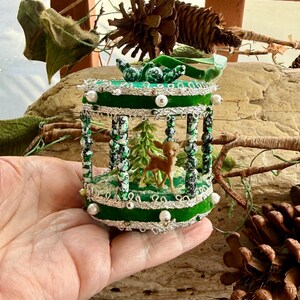 May include: A cylindrical green velvet ornament featuring a miniature brown deer figurine and a small green tree inside. The structure is supported by columns of black and white speckled beads, accented with silver trim and white pearl beads.