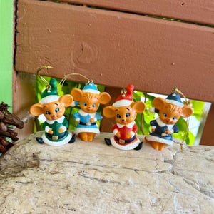 May include: Four small mouse figurines designed as holiday ornaments, each wearing a different coloured outfit including green, light blue, red, and dark blue, with matching hats and gold hanging strings attached to the top of their heads.