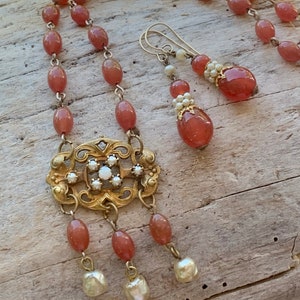 May include: A necklace and earring set featuring oval-shaped reddish-brown beads linked by metal chains. The necklace includes a central ornate gold-toned metal filigree pendant accented with small white stones and three dangling white pearl-like beads.