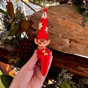 May include: A small elf figurine ornament featuring a red felt hat and matching red felt body. The hat is decorated with gold floral accents and a small pom-pom at the tip. The elf has a plastic face with painted features and a gold collar.