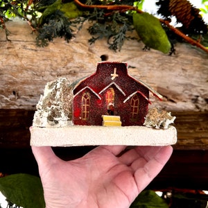 Puede incluir: Una iglesia roja en miniatura con una cruz blanca en el tejado y adornos dorados. La iglesia tiene ventanas arqueadas y está sobre una base blanca con un pequeño árbol y detalles decorativos. La iglesia mide unos 7.6 cm de largo.