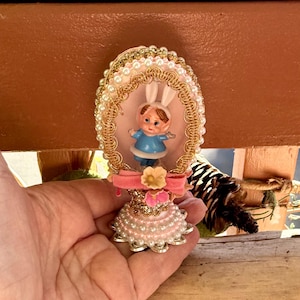 May include: An Easter-themed ornament featuring a small figurine of a child in a blue outfit with bunny ears. The ornament is egg-shaped, decorated with pink pearls, gold trim, and a pink bow. A small yellow flower is also included.