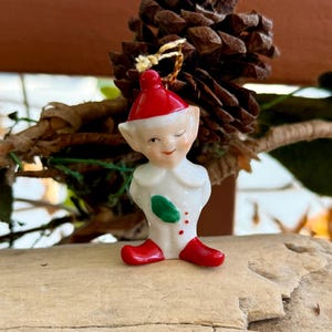 May include: A vintage Christmas ornament of a winking elf. The elf is wearing a red hat and shoes, a white outfit with a green leaf and red dots. The ornament is hanging near a pinecone.