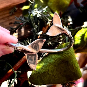 Op de afbeelding: Een zilverkleurige armband met twee gepolijste walvisstaartmotieven aan elk uiteinde van de open band. Het metaal heeft een glad, reflecterend oppervlak dat het licht opvangt en de gebogen, waaiervormige structuur van de walvisvinnen toont.