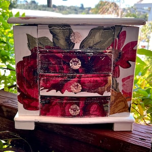 May include: A small, white jewelry box with three drawers, adorned with a floral design featuring large red roses and green leaves. The drawers have crystal knobs. The box is painted with a distressed finish, giving it a vintage look.