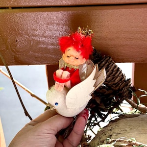 Puede incluir: Adorno navideño vintage con un pequeño querubín de pelo rojo y atuendo rojo, sentado sobre una paloma blanca con ribetes dorados. El adorno incluye una piña y detalles dorados.