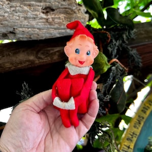 May include: A small elf figurine with a red felt hat and matching red outfit featuring a white collar and white cuffs. The elf has a moulded plastic face with painted blue eyes, rosy cheeks, and a smiling mouth, depicted in a seated position.
