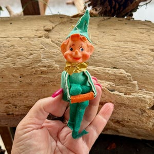 Puede incluir: Un adorno de elfo de fieltro verde con un lazo amarillo y manos de fieltro naranja. El elfo tiene una cara pintada con ojos azules y una amplia sonrisa.