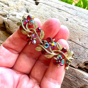 May include: A gold-toned floral brooch featuring red and iridescent crystal accents. The brooch has a branch design with leaves and small, round red crystals. The flowers have a mix of red and iridescent crystals.