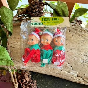 May include: A set of three vintage Pixie Doll ornaments with gold hangers, packaged in a clear plastic bag with a header card labeled Pixie Doll with Gold Hanger. Each doll features a painted face, white hat, and red or green felt clothing.