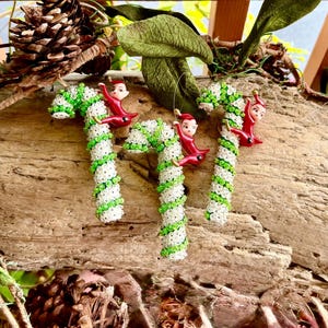 Puede incluir: Tres adornos navideños de bastones de caramelo con rayas de cuentas blancas y verdes y pequeñas figuras de elfos rojos. Los adornos cuelgan de una superficie de madera con piñas y vegetación en el fondo.