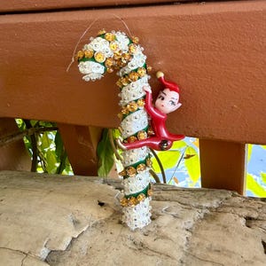 Puede incluir: Un adorno decorativo en forma de bastón de caramelo con una base en espiral verde y blanca decorada con lentejuelas transparentes y ámbar. Una pequeña figura de elfo rojo con gorro puntiagudo y rasgos pintados está sujeta al bastón.