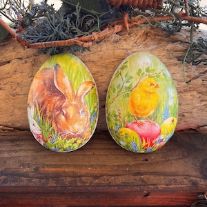 May include: Two wooden Easter egg decorations. One egg features a brown rabbit in a grassy field with blue and purple flowers. The other egg shows a yellow chick perched on a red egg, surrounded by colorful eggs.