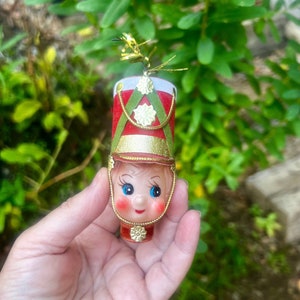 May include: A vintage Christmas ornament shaped like a toy soldier. The ornament features a red and gold hat with green accents, a painted face with blue eyes, and a red uniform. The ornament is held in a hand against a background of green foliage.
