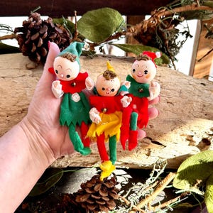 May include: Three small elf figurines with painted faces, brown hair, and felt clothing. The left elf wears green and red, the centre elf wears red and yellow, and the right elf wears red and green. Each elf features a small bell on its collar.