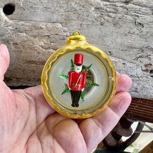 May include: A vintage Christmas bauble with a clear, yellow-edged design. Inside, a miniature toy soldier in a red coat and black trousers stands before some greenery. The bauble is held in a hand.