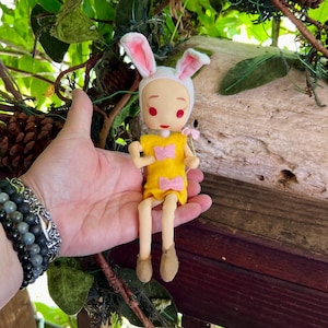 Peut inclure: Une poupée faite à la main habillée en lapin, avec un bonnet blanc et des oreilles doublées de rose. La poupée porte une robe jaune avec des nœuds roses et tient une petite fleur rose. Les jambes de la poupée sont beiges et elle a des chaussures marron.