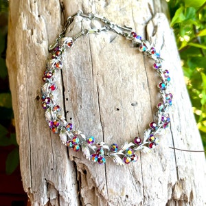May include: A silver-toned necklace with iridescent, colorful rhinestones and leaf-shaped accents. The necklace is set against a weathered wooden surface, showcasing its intricate design and vintage style.