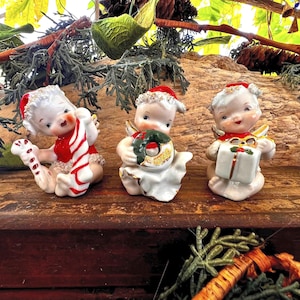 Könnte beinhalten: Drei Keramikfiguren von Kindern mit roten Weihnachtsmützen. Die erste hält eine rot-weiß gestreifte Zuckerstange, die zweite einen grünen Kranz mit goldenen Akzenten und die dritte ein weißes Geschenkpaket mit roter Schleife und grünem Blatt.