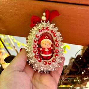 May include: An oval-shaped Christmas ornament featuring a central figurine of a child in a red and white outfit. The frame is decorated with silver metallic trim, red circular accents, and clear beads. A red fabric bow sits at the top with a pearl.