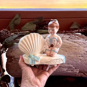 May include: A vintage porcelain figurine featuring a baby in a swimsuit, holding a small object, seated on a seashell-shaped base. The figurine is painted in soft pastel colors, with blue accents. The base has decorative scrollwork.