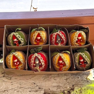 Op de afbeelding: Een set van acht decoratieve ornamenten in de vorm van appels, elk met een klein kabouterfiguurtje in een uitgesneden midden. De appels zijn gekleurd in tinten groen, geel en rood, afgewerkt met gouden kanten randen en groene stoffen blaadjes.