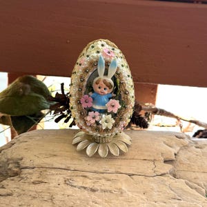 Könnte beinhalten: Ein Osterei-Ornament mit einer Figur eines Kindes mit Hasenohren und einem blauen Outfit. Das Ei ist mit Perlen, Blumen und goldenen Verzierungen verziert. Das Ornament steht auf einem goldenen Blumenfuß.