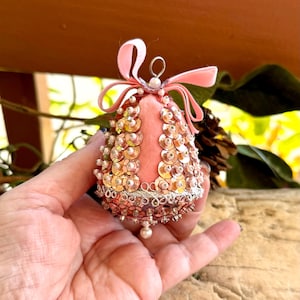 May include: A pink bell-shaped ornament with gold sequins and pearl accents. The ornament has a pink velvet bow at the top and a pearl bead at the bottom. The ornament is held in a hand.
