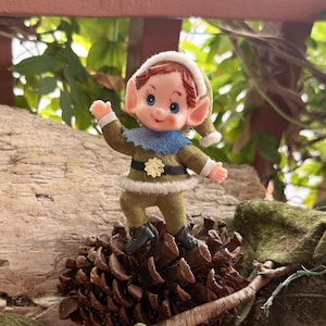 May include: A small elf figurine with a plastic face, blue eyes, and red hair, wearing a green felt outfit with white trim, a blue collar, and a gold star-shaped belt buckle. The elf has pointed ears and is posed with one arm raised in a wave.