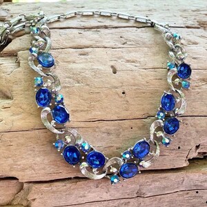 Puede incluir: Un collar de metal plateado que presenta una serie de piedras ovaladas de color azul intenso facetadas y cristales de acento azules iridiscentes más pequeños. Los eslabones metálicos tienen un acabado cepillado y un patrón curvo.