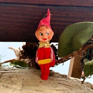 Könnte beinhalten: Ein Vintage-Weihnachtselfen-Ornament. Der Elf trägt einen roten Filzanzug und -hut, eine goldene Fliege und ein lächelndes Gesicht mit blauen Augen. Der Schmuck hängt an einer goldenen Schnur.