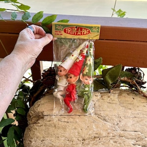 Könnte beinhalten: Ein Set aus drei Vintage-Wichtel-Anhängern in einer durchsichtigen Plastikverpackung mit einem Pappschild mit der Aufschrift TRIM-A-PAC. Die Wichtel tragen spitze Mützen und Outfits in Weiß, Rot und Grün mit kleinen Blumen.