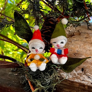 May include: Two vintage felt Christmas ornaments shaped like snowmen. The snowman on the left is wearing a yellow and red hat and a yellow dress. The snowman on the right is wearing a green hat and a red and blue dress.